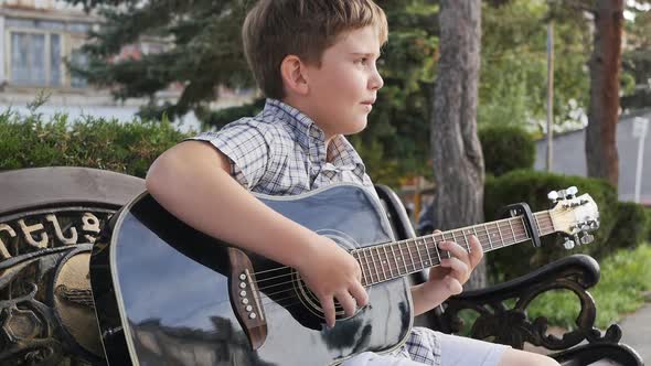 Portrait a Boy of School Age Sitting in a Park on a Bench Plays a Black Acoustic Guitar with a White
