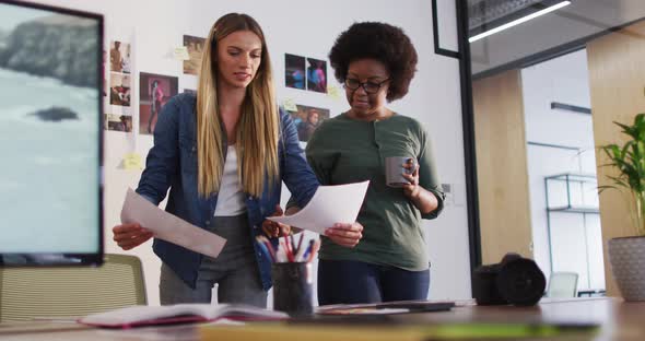 Two diverse female colleagues discussing and looking at images standing in office alt