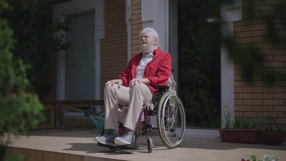 Wide Shot of Old Disabled Caucasian Man in Wheelchair Enjoying Sunny Summer Day Outdoors on House alt