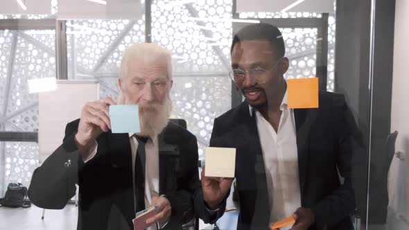 Motivated Managers Standing Near Glass Window Wall with Colorful Stickers Discussing Project alt