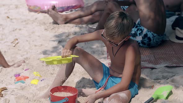 Boy in Blue Swimming Trunks Sows Sand Through Sieve on Beach alt