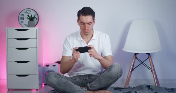 Young Man is Sitting on the Floor at Home and Playing Game Using His Smartphone alt