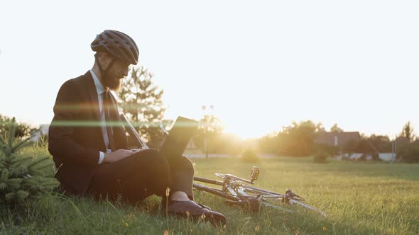 Man Sitting on a Grass at the Park Working on a Laptop Wearing a Helmet alt