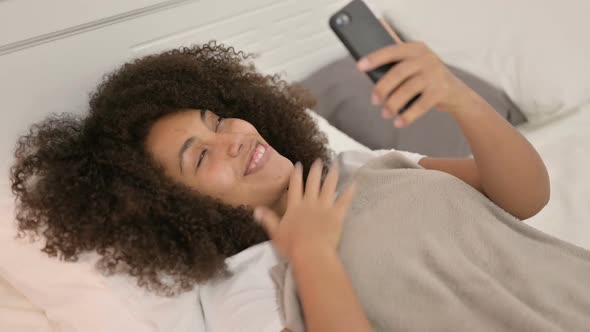 Video Chat on Smartphone By Young African Woman Laying in Bed alt