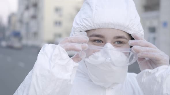 Close-up Face of Smiling Young Woman in Face Mask and Antiviral Suit Putting on Protective alt