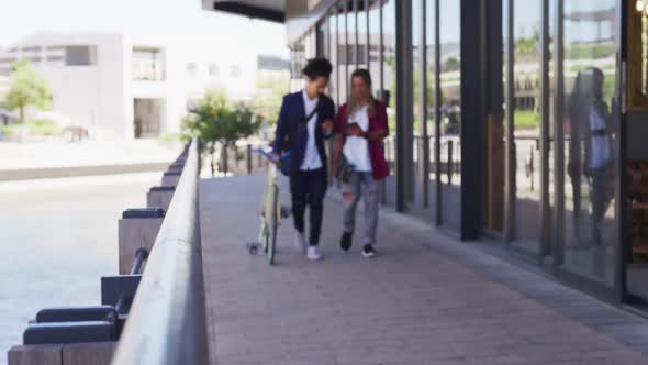 Two diverse male friends walking in the street using a digital tablet and wheeling a bicycle alt