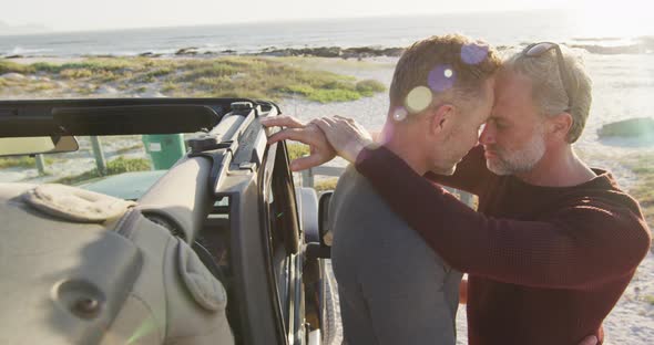 Happy caucasian gay male couple standing by car embracing on sunny day at the beach alt