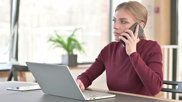 Young Woman with Laptop Talking on Smartphone in Office  alt