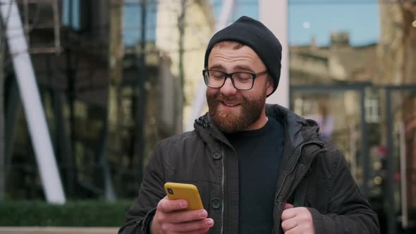 Close Up View of Bearded Male Person in 30s Looking at Phone Screen and Smiling While Walking at alt