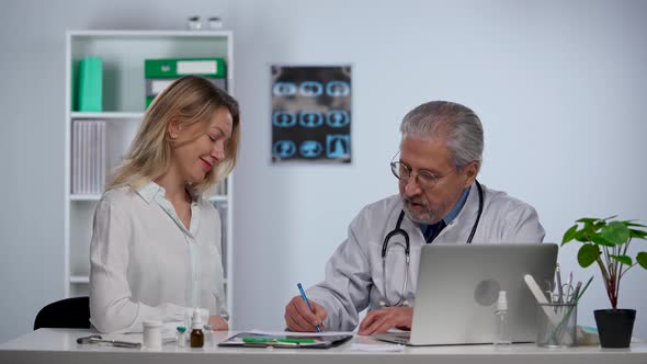 Man Doctor in White Medical Coat Sits at Table in Clinic Looks at Snapshot of Patient and Speaking alt