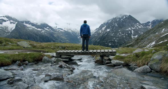 Hiker Standing on River Bridge in Front of Alpine Cloudy Mountains Enjoying Nature alt