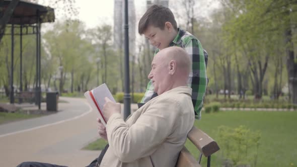 Portrait Cute Grandfather and Adorable Grandson Sitting in the Park on the Bench alt