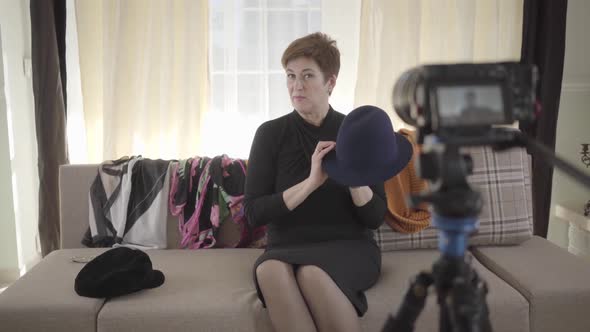 Portrait of Positive Senior Caucasian Woman Showing at Camera Hats and Scarfs. Mature Woman Sitting alt