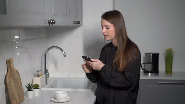 A Young Woman in the Kitchen Uses a Mobile Phone