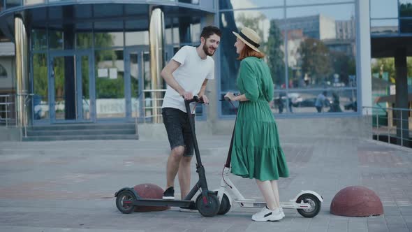 Young Couple in Love Having Fun with Kisses During Walking on Electric Scooters alt