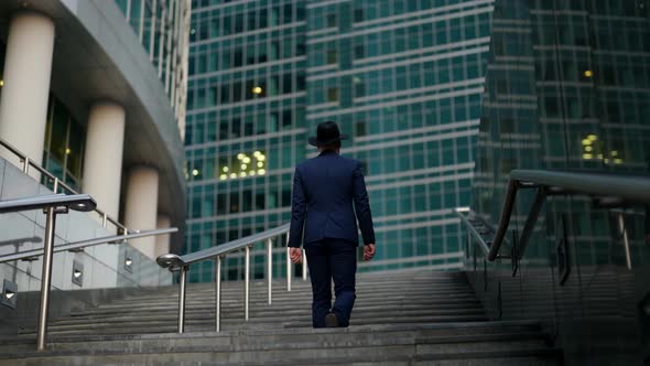 a Stylish Man in a Plaid Suit and Hat Climbs the Stairs Against the Background of a Blurry Modern alt