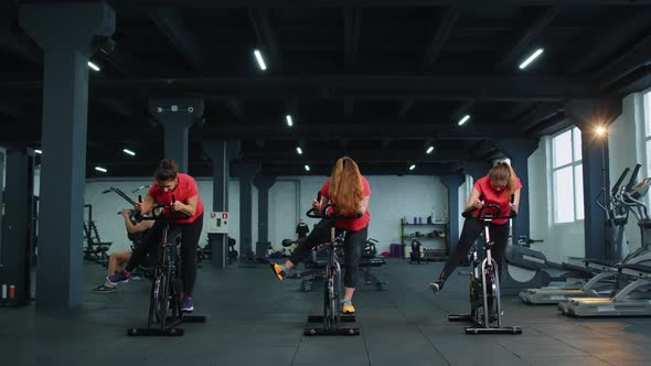 Group Athletic Girls Performing Aerobic Riding Training Exercises on Cycling Stationary Bike in Gym alt