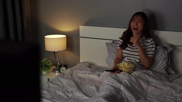 funny young woman watching TV and laughing on a bed at night