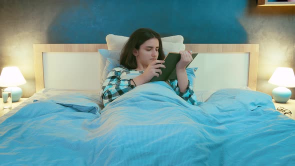 Teenage Girl Lying in Her Bed in the Evening Reading a Book Front View alt