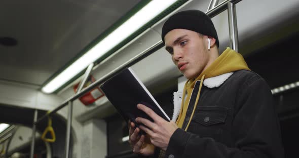 Side View of Young Man with Wireless Earpods Turning Pages While Enjoing Story. Millennial Handsome alt
