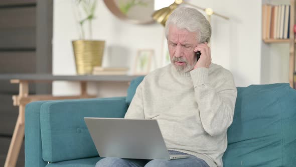 Old Man with Laptop Talking on Smartphone on Sofa  alt