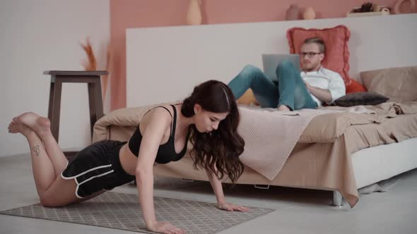Woman Doing Push Ups at Home While Boyfriend Man Sits on Bed and Uses Laptop. Fit Female Doing Sport