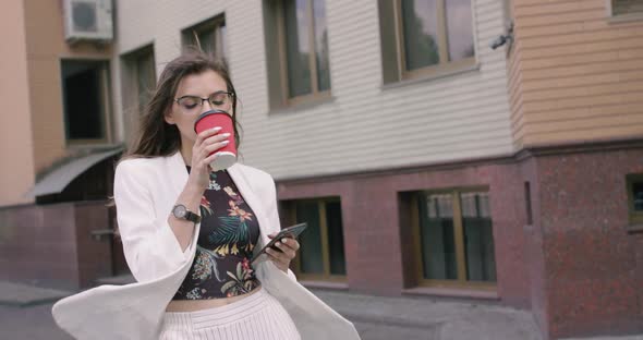 Lady with Blowing Hair and Jacket From Wind Drinks Coffee, Uses Phone and Smiles alt