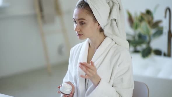 Young Woman in White Bathrobe and with Towel on Head Applies Moisturizer Cream on Her Face alt