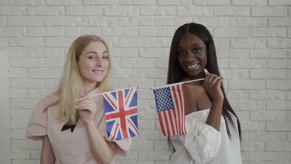 Young Women Holding British and American Flags Smiling at Each Other. Caucasian and African American alt