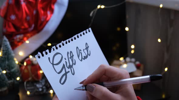 Female Hand Writing Gift List Sheet of Paper with Text GIFT LIST Against Christmas Decorations alt