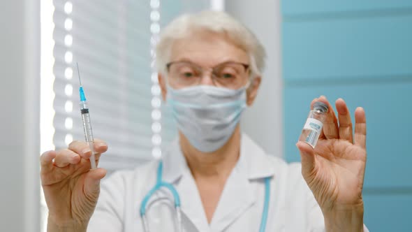 Mature woman doctor with spectacles and disposable mask shows vial of vaccine alt
