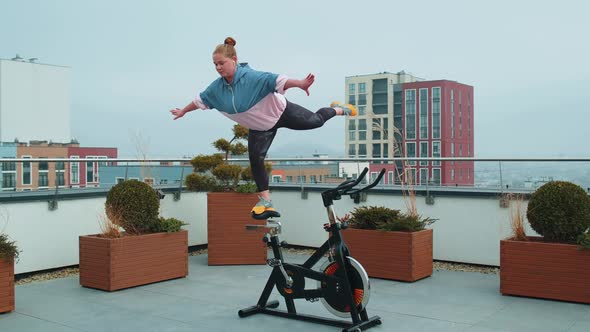 Athletic Girl Performing Swallow Acrobatic Trick Exercises on Cycling Stationary Bike on House Roof alt