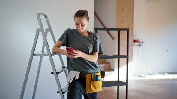 A Professional Repairwoman Uses Her Phone While Leaning on a Stepladder
