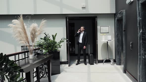 Serious Businessman with Suitcase is Talking on Mobile Phone in Hotel Hall alt