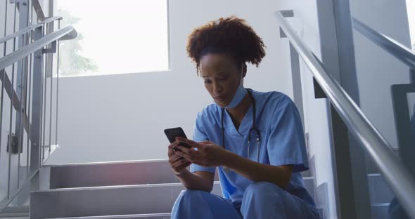Mixed race female doctor wearing face mask sitting on stairs in hospital using smartphone alt