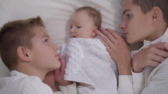 High Angle View Charming Adorable Infant Girl Lying on Bed with Teenage Boys Admiring Child alt