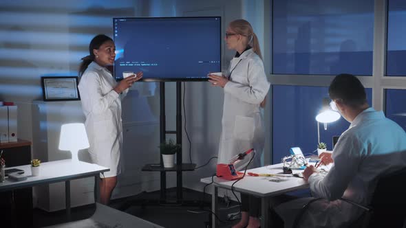 Female Engineers Talking About Something Near the TV Screen with Cup of Drink alt