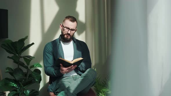 Bearded Young Man Sitting Near Window and Reading a Book at the Sunrise alt