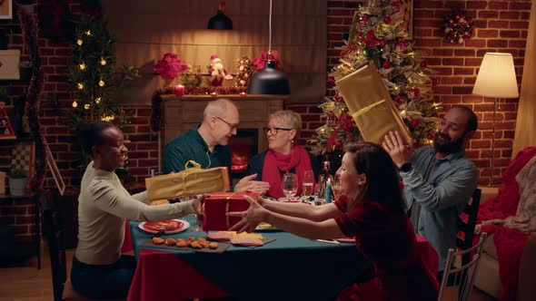 Joyful People Gathered at Christmas Dinner Table While Exchanging Gifts alt