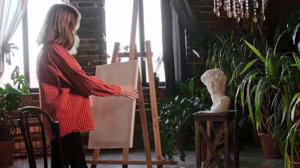 A Young Pretty Woman Artist Putting a Canvas on an Easel and Sits in Front of It alt