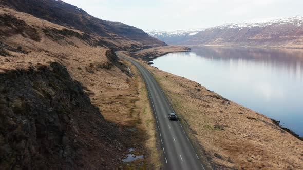 Drone Tracking Car Driving By Fjord And Towards Mountains alt