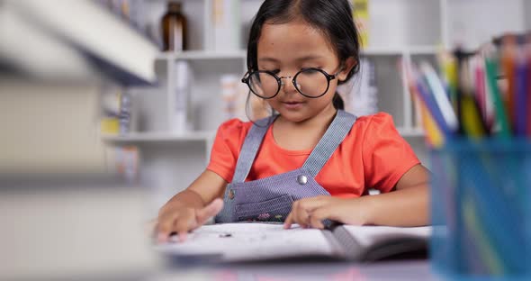 Asian little girl wearing glasses practicing reading at the desk at classroom. alt