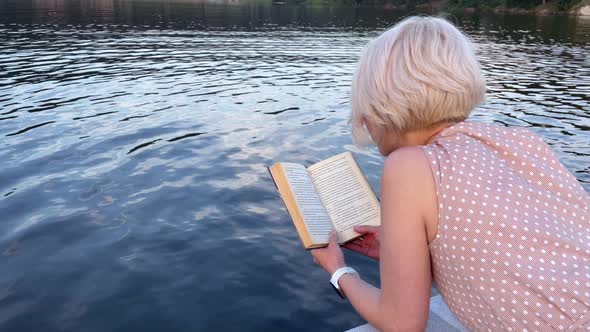 A Blonde with Short Hair Reads a Book Against the Background of a Lake