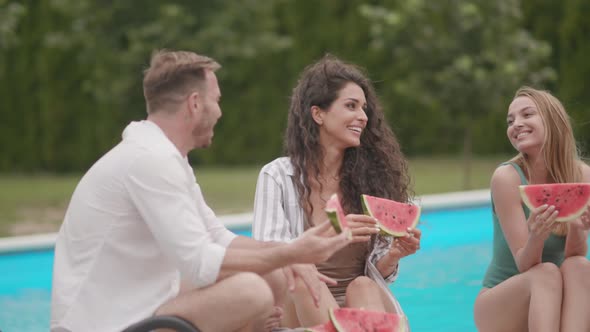 Group of young people sitting by the swimming pool and eating watermelon in the house backyard