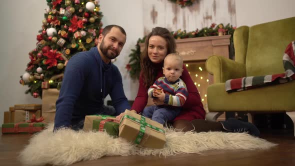Happy Family Celebrating Christmas Together Mother, Father and Little Baby Sitting on the Floor alt