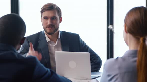 Two businessmen handshake and smiling while sitting at a table with a team alt