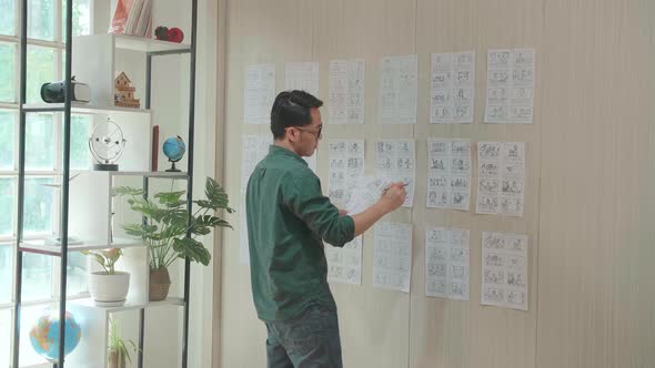 Young Creative Asian Designer Looks At The Storyboard Sketches Covering His Wall alt