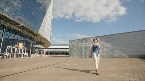 Pretty Young Girl Walking In Highheels In A Long Light Dress Along The Huge Glass Building  alt
