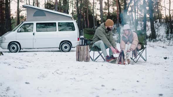 Couple is Sitting By a Campfire in the Woods in Winter Enjoying Van Life