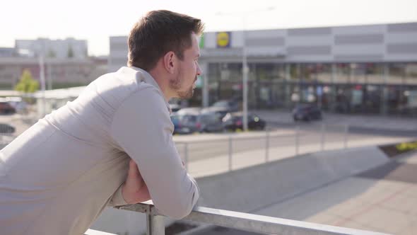 A Young Caucasian Man Looks Around on an Overbridge in an Urban Area  Parking Lot and a Supermarket alt